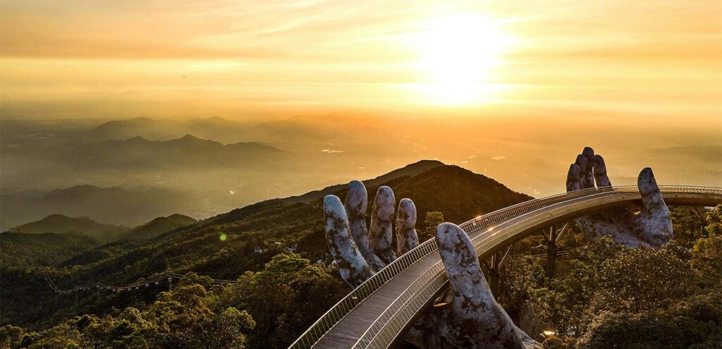 A dramatic sunrise view of the Golden Bridge in Ba Na Hills, Da Nang. The curved pedestrian walkway is supported by two massive, weathered stone hands that appear to lift it high above the mist-shrouded green mountain valleys.
