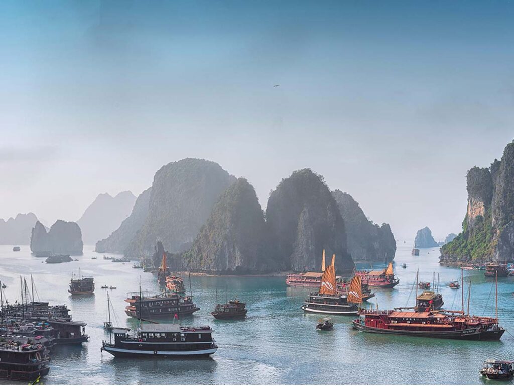 A high-angle, scenic view of Ha Long Bay in Vietnam. Its is one of the best things to do in Vietnam. Numerous traditional wooden junk boats and cruise ships float across the calm, greenish-blue water. In the background, massive, steep limestone karsts covered in green vegetation rise out of the sea, fading into a hazy, light blue horizon.