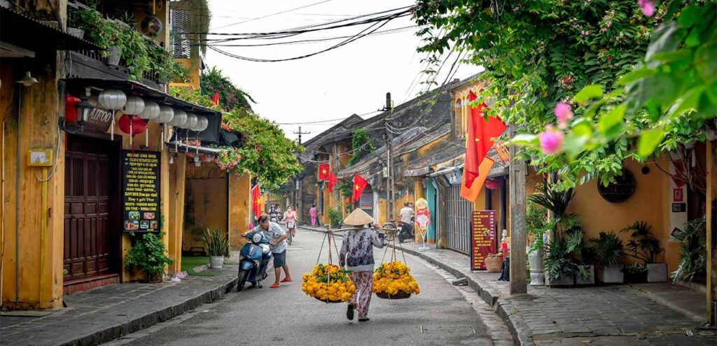 A street scene featuring iconic yellow-painted historic buildings with a street vendor in the foreground carrying baskets of yellow flowers on a traditional wooden shoulder pole. (Note: While the filename says Hanoi, this distinctly depicts the classic architectural style of Hoi An Ancient Town).