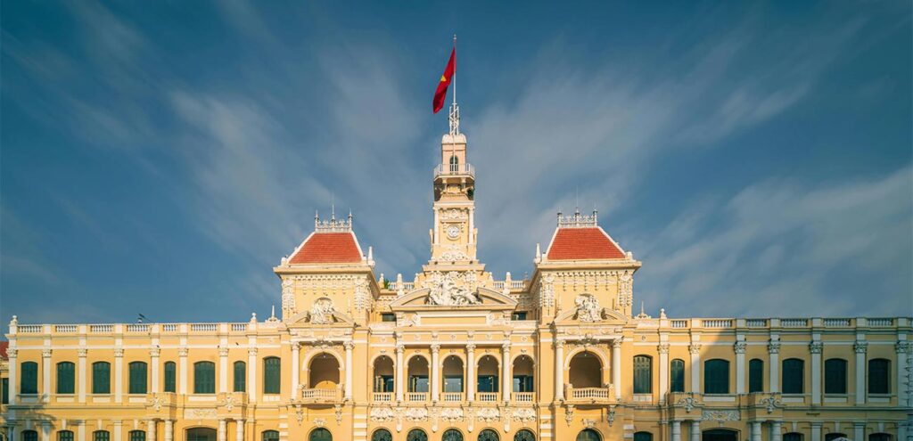 The symmetrical front facade of the historic Ho Chi Minh City Hall. The French colonial-style building features elegant yellow and white architecture, ornate pillars, and the red and yellow flag of Vietnam flying high from its central clock tower against a blue sky.