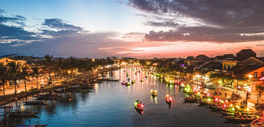 An elevated evening view of the Thu Bon River in Hoi An Ancient Town. The river and its banks are beautifully illuminated by the warm glow of lanterns and numerous small tourist boats, set against a dramatic twilight sky with orange and purple clouds.