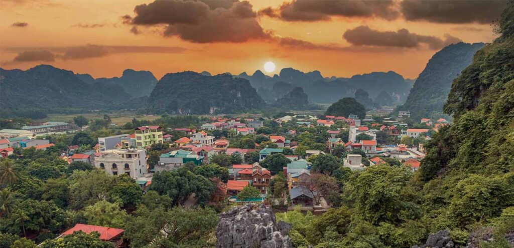 Best things to do in Vietnam is to watch a panoramic sunset view over a valley town in Ninh Binh, Vietnam. The sun glows brightly directly behind a backdrop of jagged karst mountains, casting warm orange and golden light across the clouds and the dense cluster of colorful buildings below.