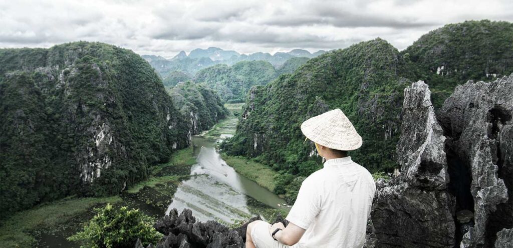 A high-angle shot from a jagged rocky peak showing a person from behind wearing a traditional Vietnamese conical hat and white clothing. They are looking out over a winding river that snakes through a vast valley of lush green karst mountains in Ninh Binh under an overcast sky.