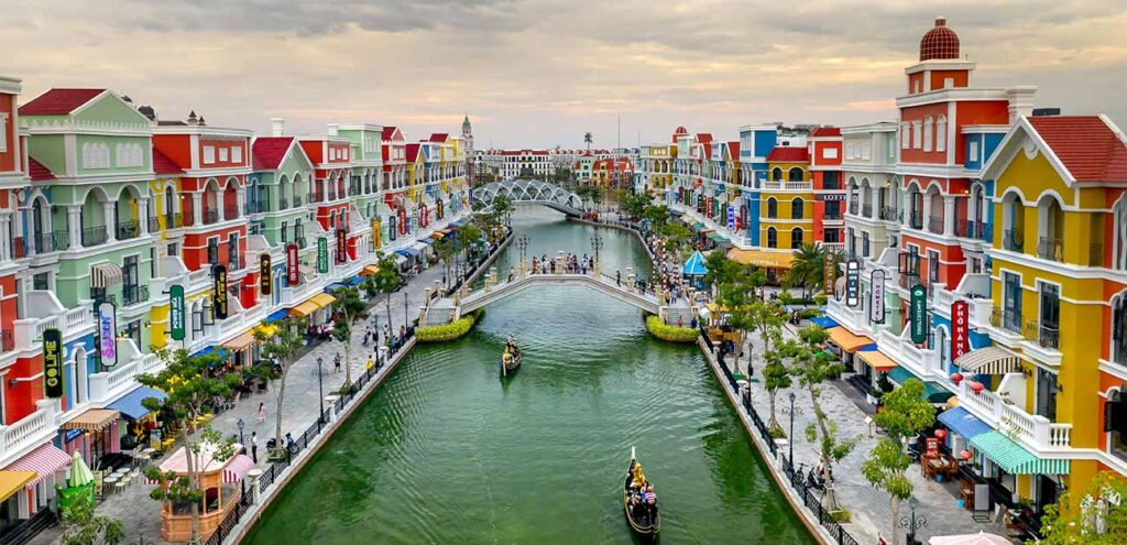 A vibrant canal scene in Phu Quoc, lined with brightly colored, European-style buildings and elegant arched pedestrian bridges. Traditional gondolas navigate the green water under a soft twilight sky, mimicking a Venice-like atmosphere.