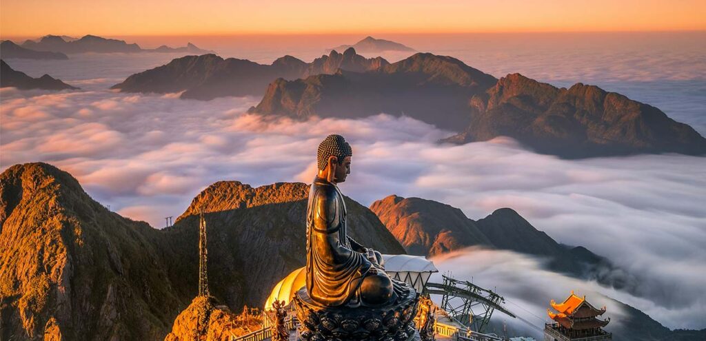 A high-angle view of a large bronze Buddha statue sitting in meditation on a rugged mountain peak in Sapa (Fansipan). The statue is captured from a side profile, looking out over a spectacular sea of thick white clouds that blankets the valleys, with sharp mountain ridges bathed in the warm, golden light of a vibrant sunset.