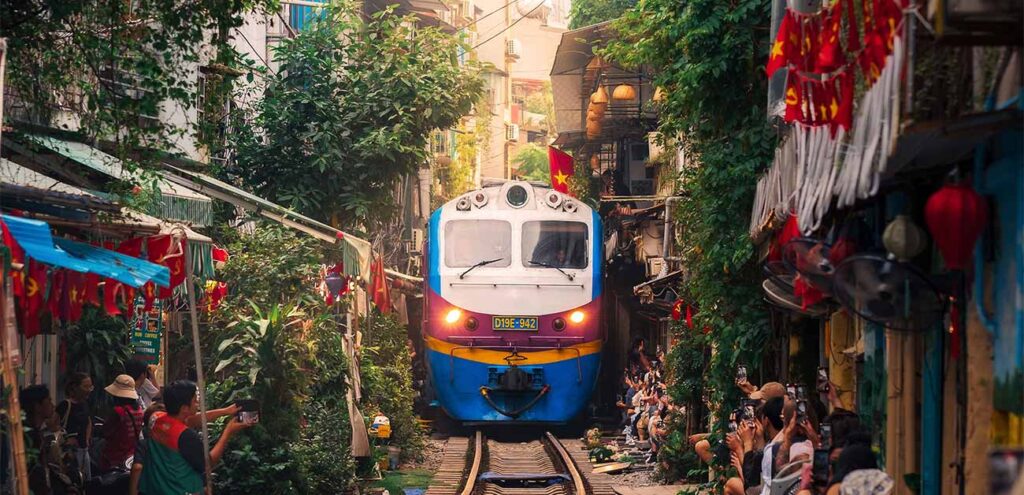 A blue and yellow train passing through the famously narrow Train Street in Hanoi, Vietnam. Crowds of tourists line the close-quarters alleyway on both sides, holding up smartphones to capture the train as it squeezes past cafes and lush green plants.