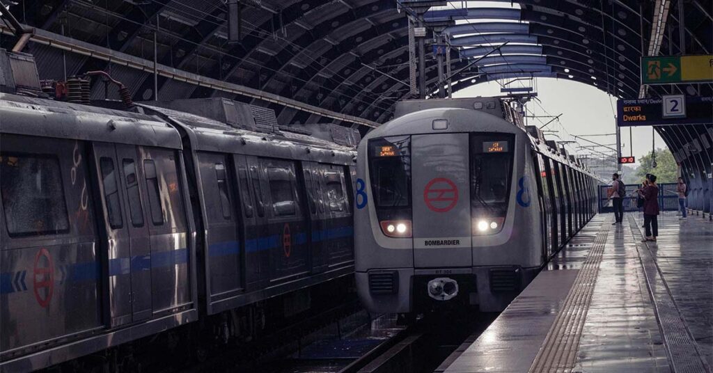 Two metro trains stand at a covered station platform, with one train facing the camera under arched metal roofing while a few passengers wait nearby.