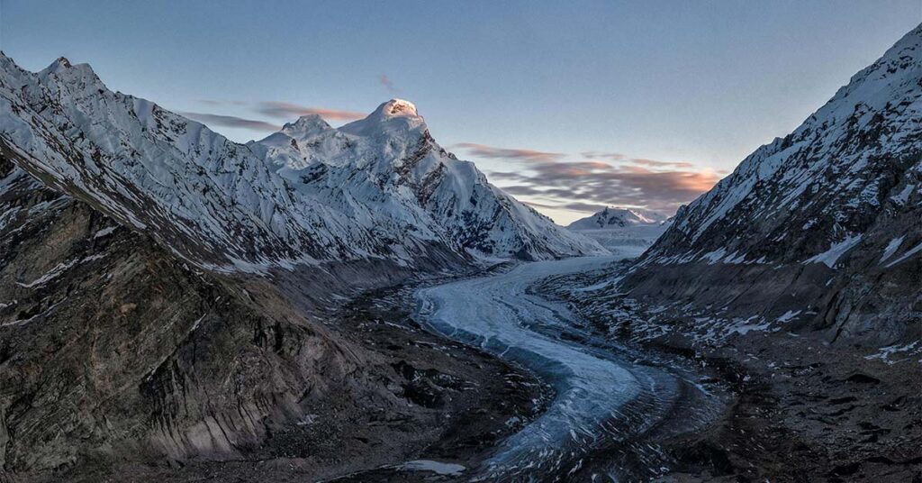Panoramic view of the frozen Drang Drung Glacier in Zanskar Valley flowing between snow-clad Himalayan mountains during evening.