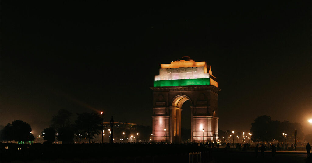 India Gate illuminated in the colours of the Indian flag at night, surrounded by streetlights and silhouetted visitors against a dark sky.