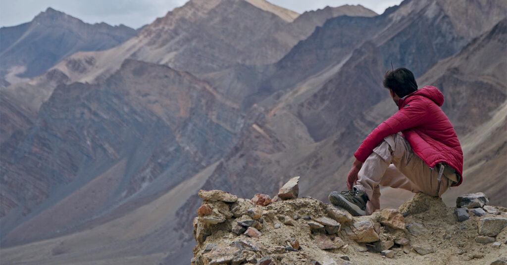 A person in a red puffer jacket sits on a rocky mountain ledge in Zanskar Valley, looking out over a rugged range of gray and brown peaks under soft daylight.