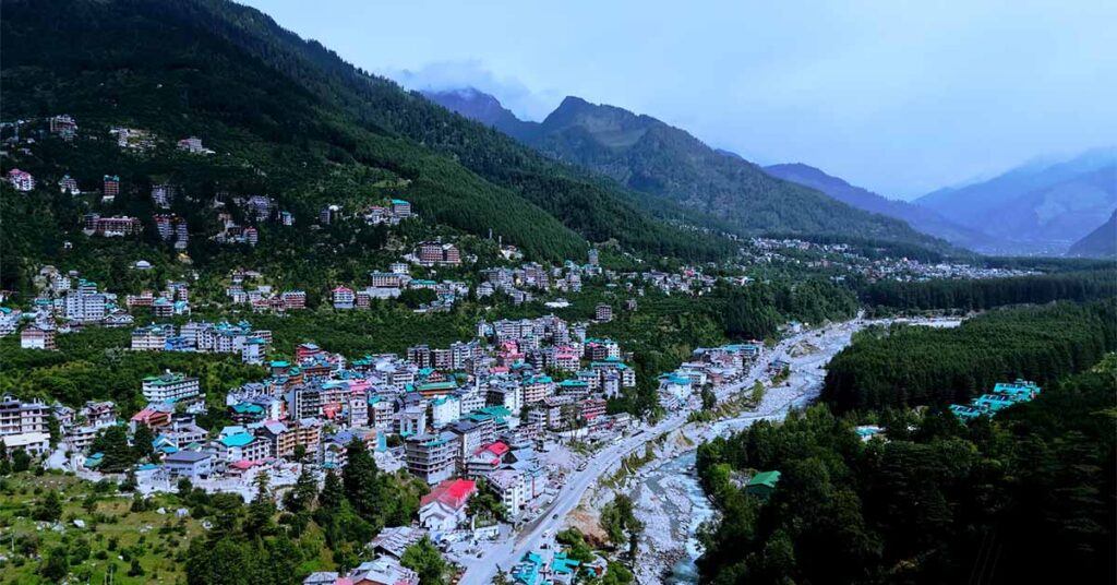 A mountain town spread across a green valley, with colorful buildings clustered along a river and forested hills rising into misty mountains in the background.