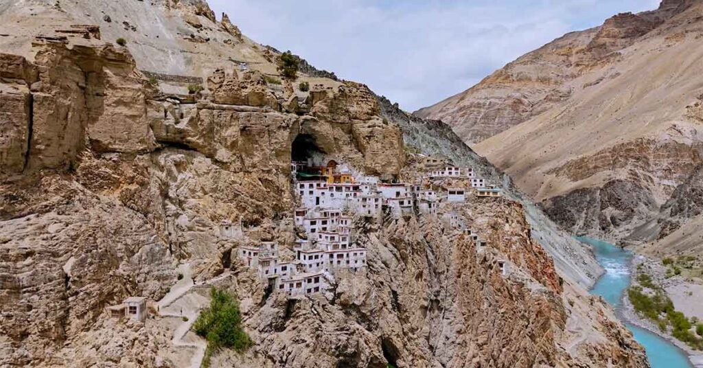A remote Phugtal monastery in Zanskar Valley complex built into steep rocky cliffs beside a turquoise river in a barren mountain valley, with white buildings clustered around a cave-like opening high on the mountainside.