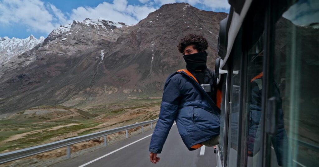 A person in a blue puffer jacket leans out of a moving bus window on a mountain road, with rugged snow-capped peaks and a cloudy sky in the background.