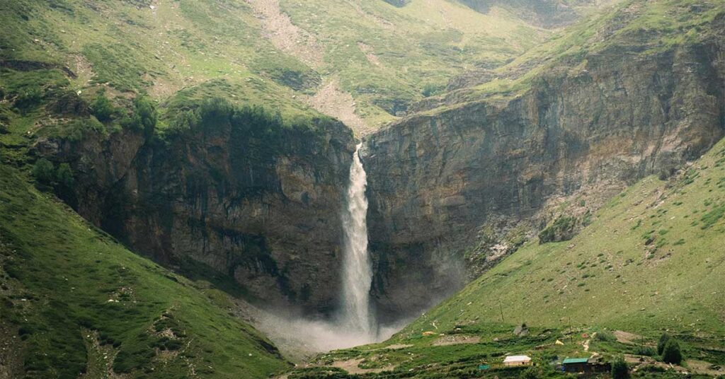 A tall waterfall plunges down a rocky cliff into a misty basin surrounded by green hills and steep canyon walls, with a few small buildings visible in the grassy valley below.