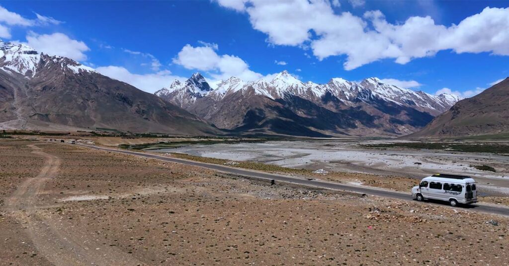 Tempo traveller driving through the vast barren landscapes of Zanskar Valley with snow-capped Himalayan ranges in the background.