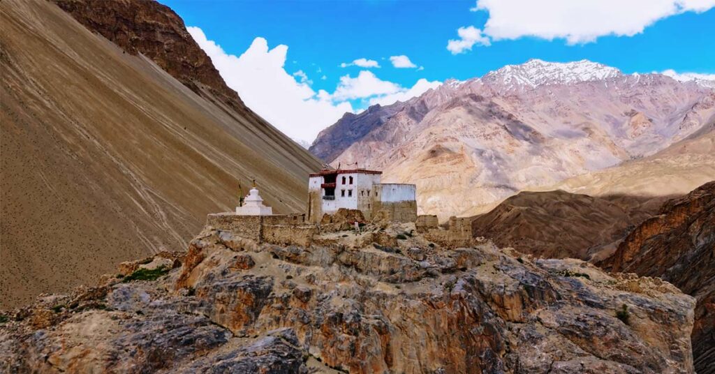 Aerial view of Zangla Palace perched on rocky cliffs in Zanskar Valley, surrounded by barren Himalayan mountains under a bright blue sky.