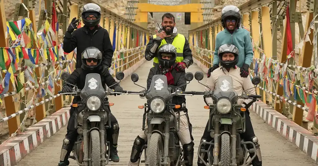 Adventure motorcyclists posing on the high Chicham Bridge decorated with prayer flags during their Spiti bike trip.