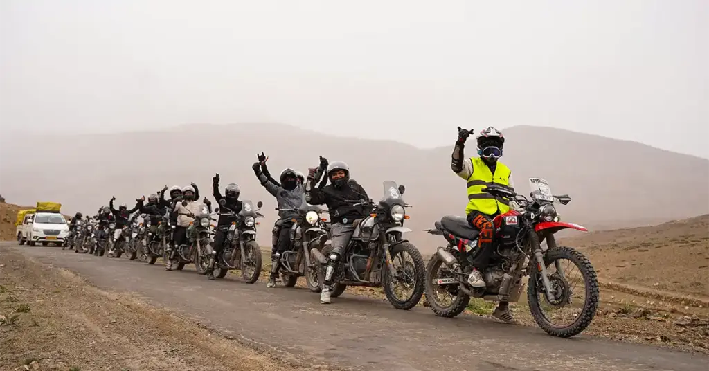 Large group of bikers on a misty Spiti Valley road with trip captain on dirt bike at front, backup vehicle visible, convoy stretching behind in cold desert terrain