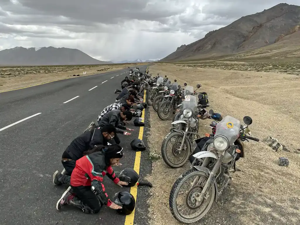 Group of bikers bowing on a well-paved Ladakh highway with Royal Enfield motorcycles parked roadside, open cold desert valley stretching ahead