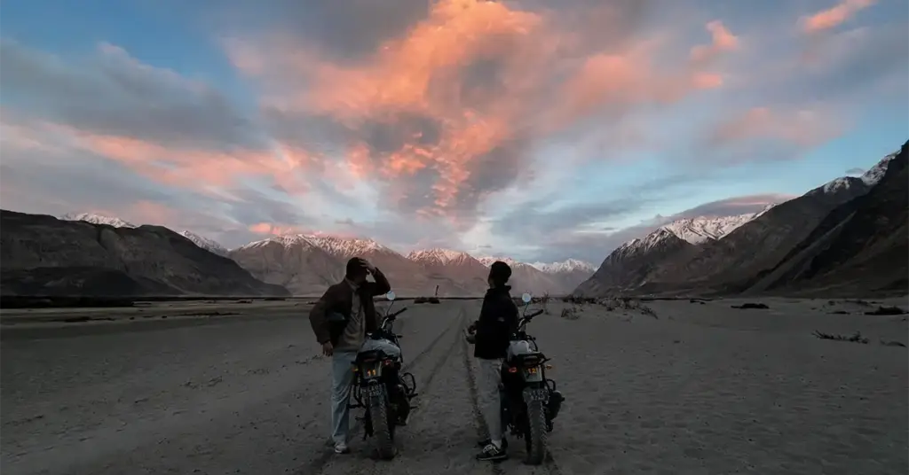 Two riders with motorcycles at sunset in Nubra Valley, Ladakh, with dramatic orange and pink clouds over snow-capped Karakoram peaks and sandy valley floor