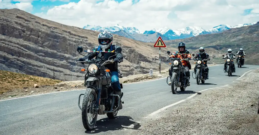 Motorcyclists wearing protective gears and riding on a paved winding highway with snow-capped mountains during an adventurous Spiti bike trip in Himachal Pradesh.