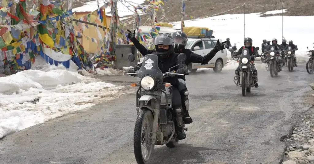 Riders crossing Kunzum Pass at 4551m in Spiti Valley with colourful prayer flags overhead, snow banks on both sides, pillion passenger raising arms in celebration