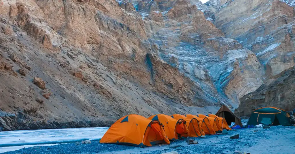 A row of bright orange expedition tents pitched on the rocky banks of the frozen Zanskar River during the winter Chadar Trek, surrounded by immense canyon cliffs in Ladakh, India.
