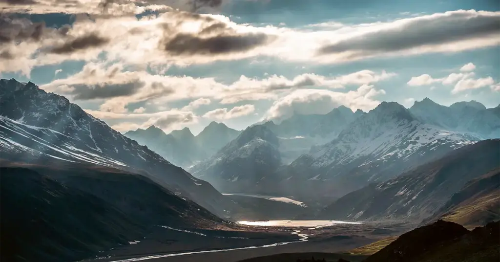 Dramatic sunbeams breaking through clouds over a winding river valley and glacial lake surrounded by towering, snow-capped Himalayan peaks in Spiti Valley.