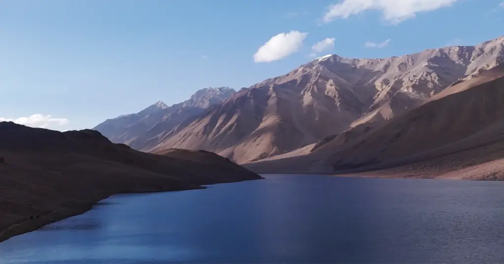 A wide, scenic landscape of a serene high-altitude lake with calm, deep blue water nestled against massive, barren, sunlit mountain ranges under a light blue sky with scattered clouds.