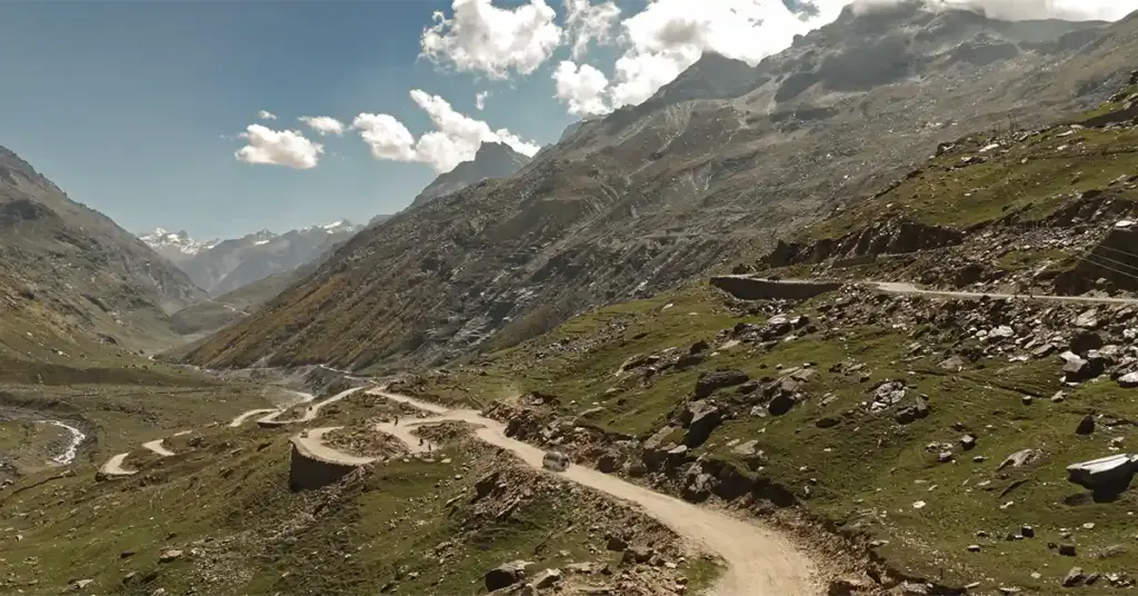 A white vehicle drives along winding mountain switchbacks on a rugged dirt road traversing a steep, rocky valley with distant snow-capped peaks.