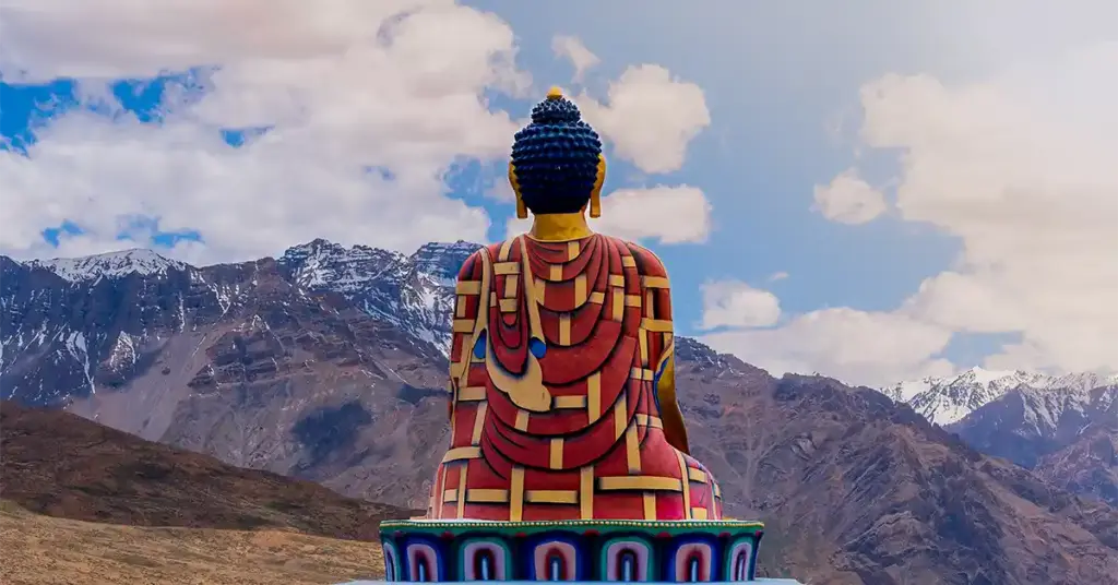 Rear view of the iconic colorful seated Buddha statue in Langza village, looking out toward the massive, rugged mountain ranges of Spiti Valley under a blue sky.