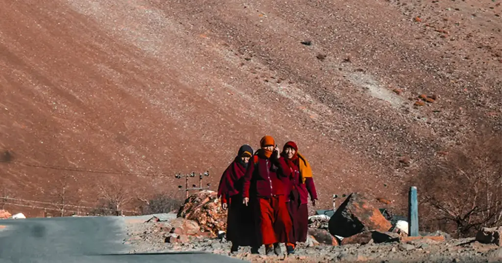 Three Buddhist monks in traditional maroon robes walking on a Spiti Valley road with rust-red cold desert mountain terrain behind them