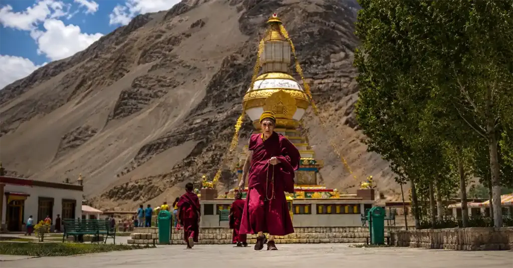 An elderly Buddhist monk in traditional maroon robes walks along a stone pathway in front of a large golden and white stupa, with towering, barren mountain slopes under a partly cloudy sky.