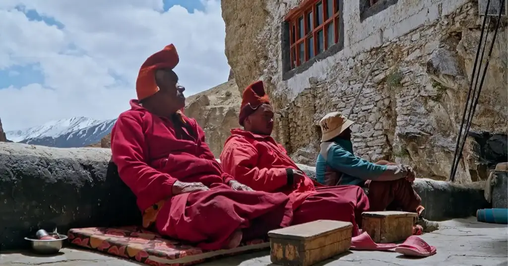 Tibetan Buddhist monks wearing traditional red robes and ceremonial hats sitting outside a stone monastery building with snow-capped Himalayan mountains in the background of Zanskar Valley, Ladakh, India.