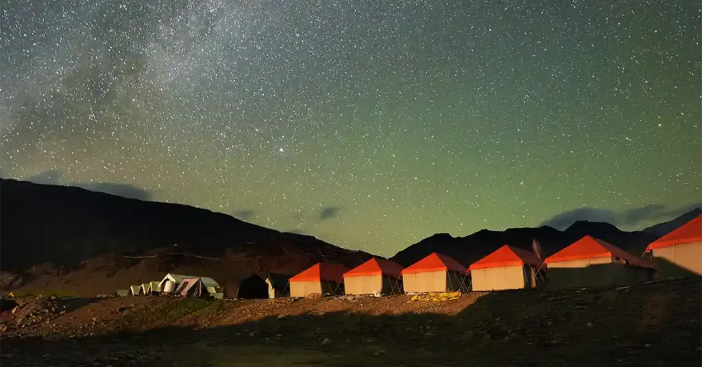 A row of illuminated orange camping tents pitched against a dark mountain silhouette under a vibrant, star-filled night sky showcasing the Milky Way galaxy.