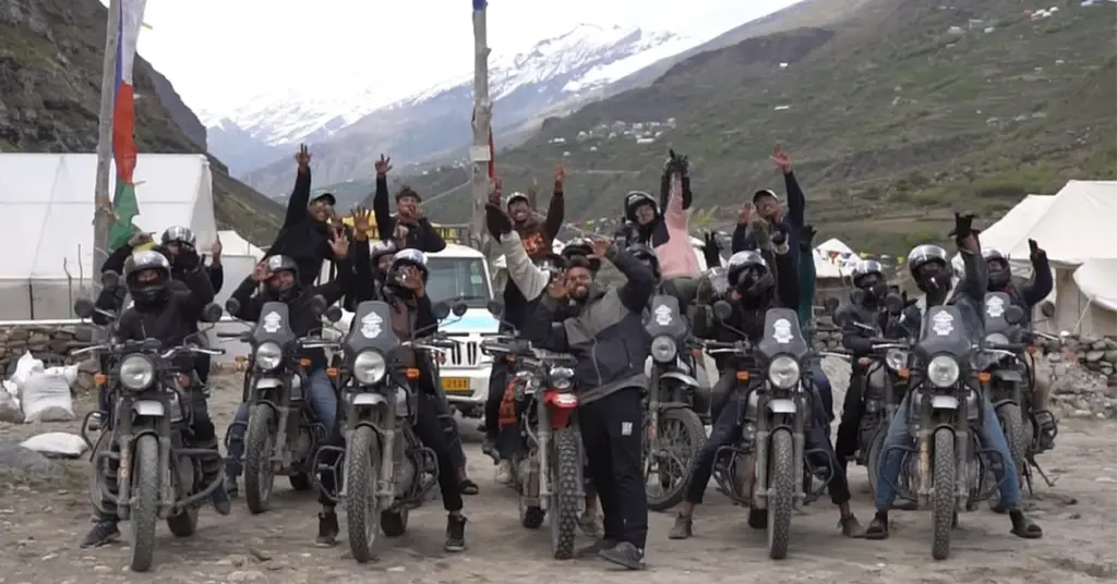 Bikers celebrating with arms raised at a Spiti Valley expedition campsite with snow-capped Himalayan peaks and prayer flags in the background