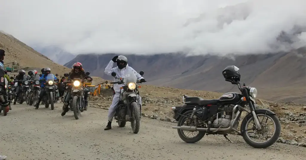 Convoy of motorcycles navigating a rough gravel road in Ladakh under heavy cloud cover with barren high-altitude mountains on both sides