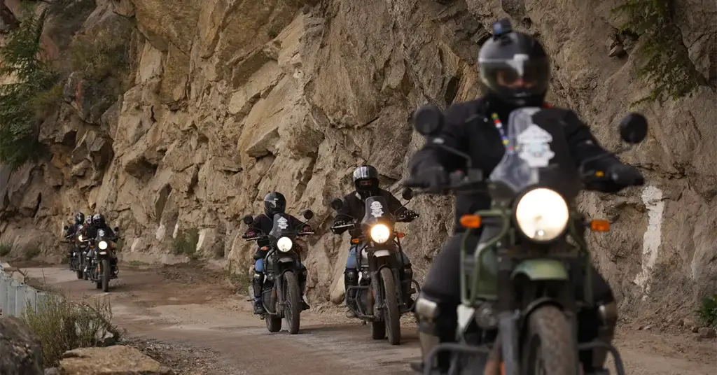 A convoy of adventure motorcyclists navigating a rugged dirt road beneath massive cliffs during a Spiti bike trip.