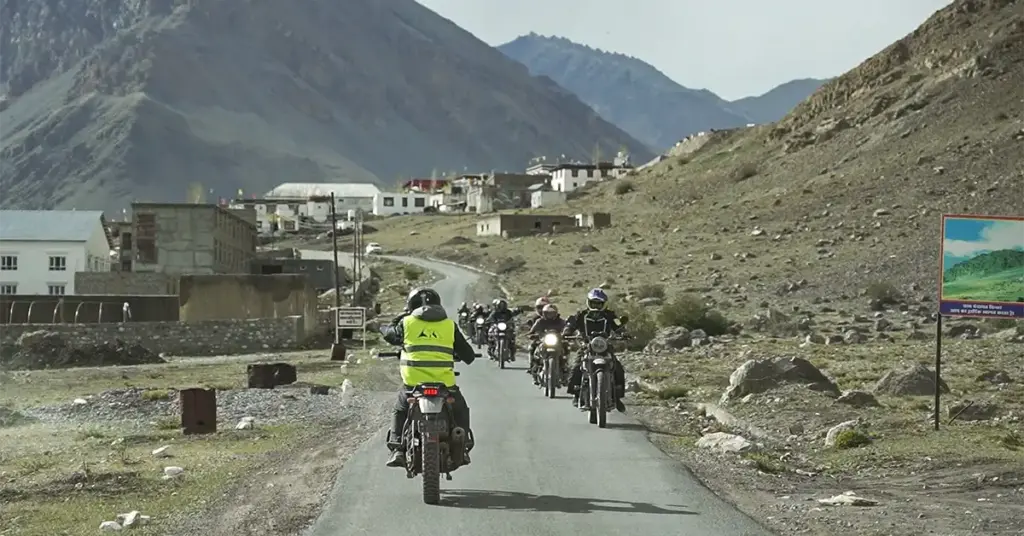 Group of bikers led by trip captain in high-visibility vest riding into a traditional Spiti village on a narrow mountain road with whitewashed buildings visible