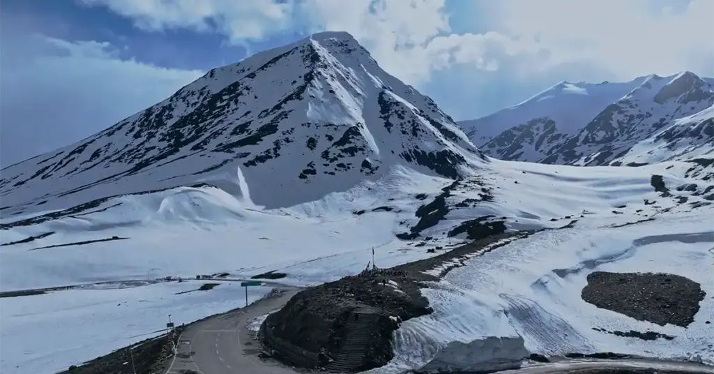 High-altitude aerial view of a winding asphalt road cutting through the heavy winter snowpack at Shinku La Pass (Shinkula Top), the mountain gateway connecting Himachal Pradesh to Zanskar Valley, Ladakh.