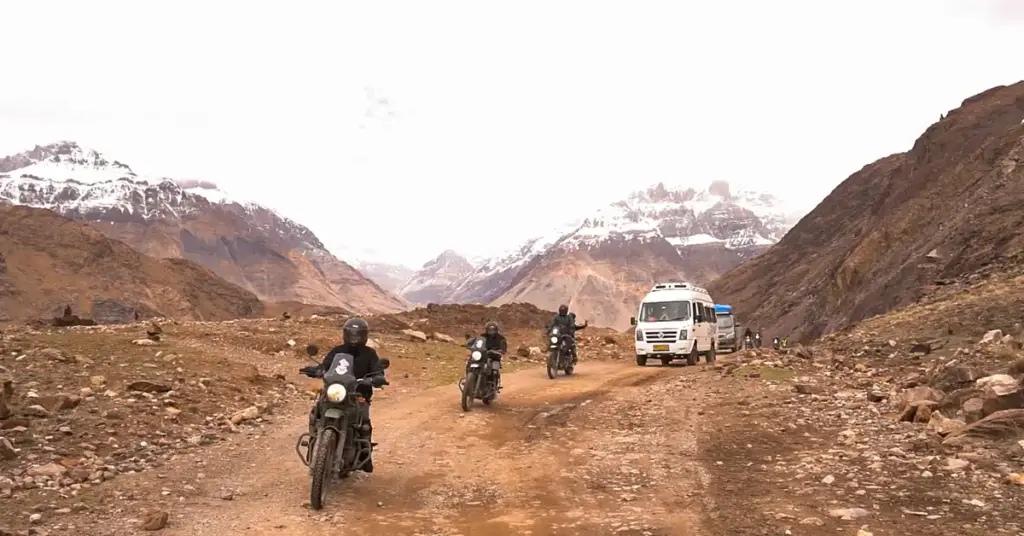 Three riders on unpaved rocky Spiti Valley track with backup tempo traveller, no road markings, snow-capped peaks ahead — typical terrain on the Losar-Chandratal route. 