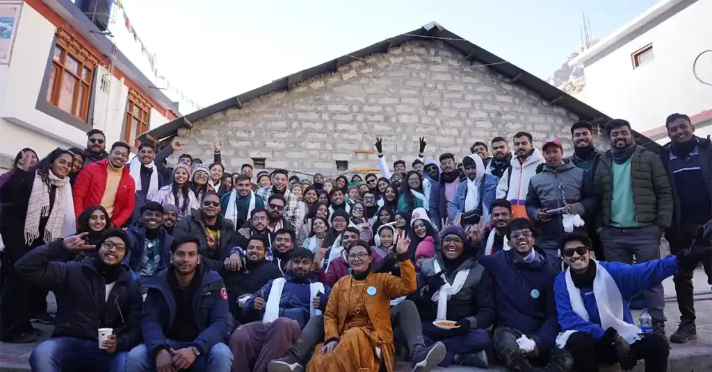 A large group of cheerful travellers posing together for a group photo in front of a traditional stone building during a winter tour in Spiti Valley.