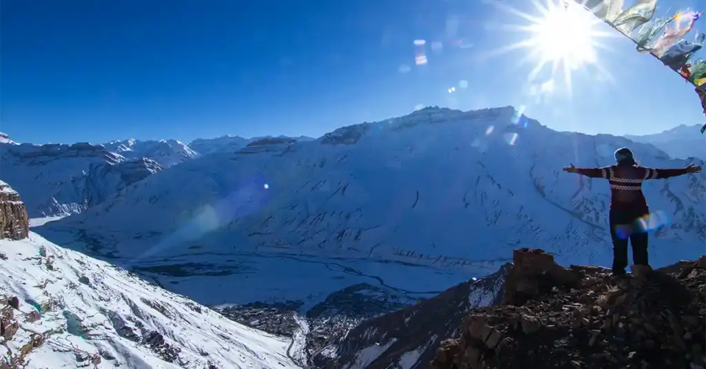 A traveler standing with open arms on a rocky cliff edge overlooking a vast, snow-covered mountain landscape under a bright winter sun in Spiti Valley.ft